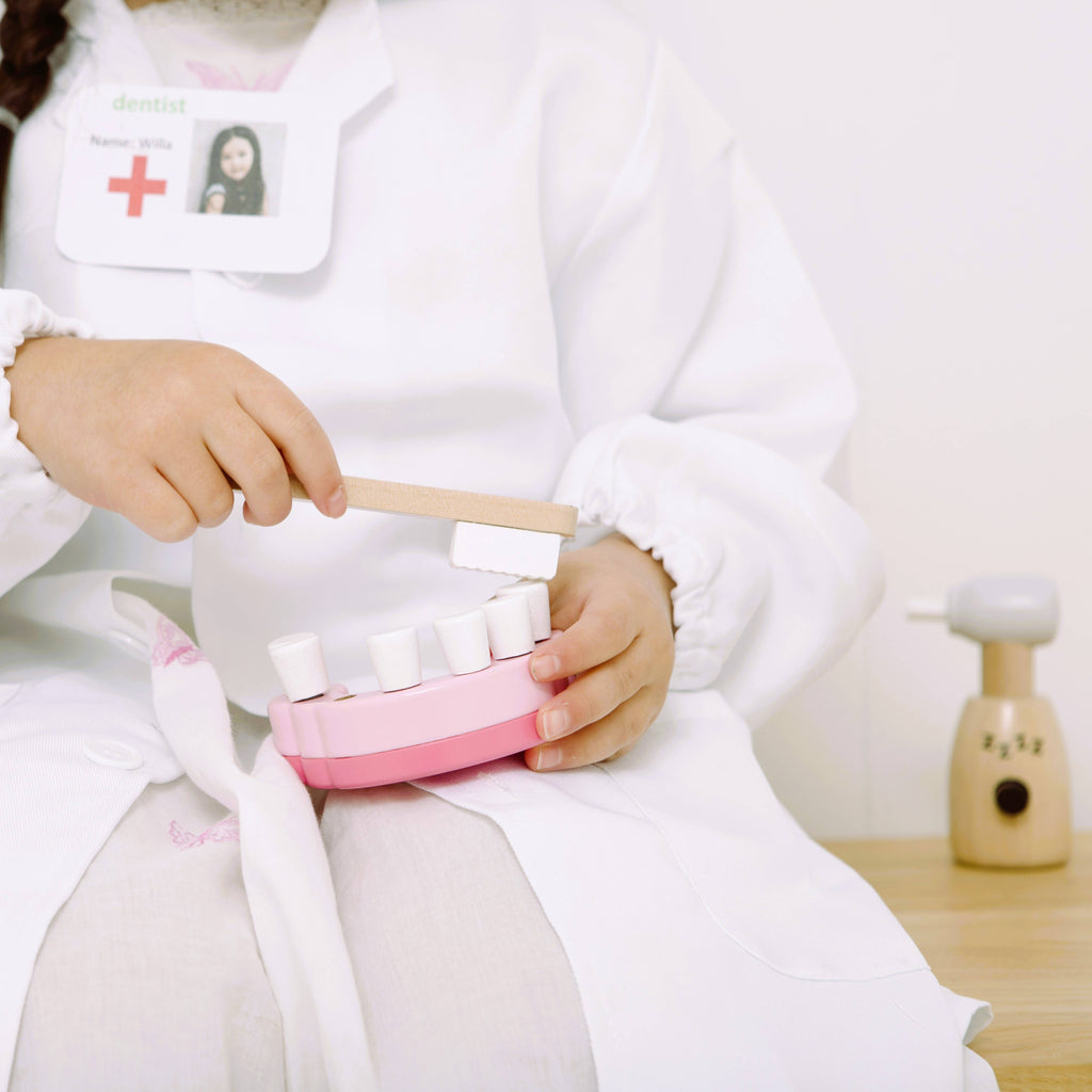 A child dressed as a dentist holds a teeth model and uses a toy toothbrush, with the Wooden Dentist Set’s wooden dental tool from their role play kit visible on the bench nearby.