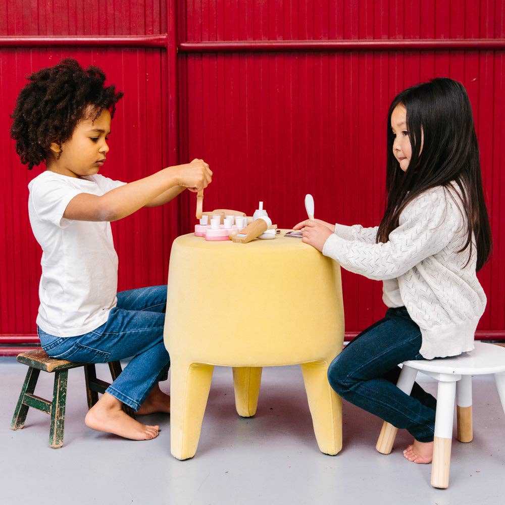 Two young children sit on small stools at a yellow table, playing with a Wooden Dentist Set. The background is a red wall. Both children are barefoot and appear focused on their imaginative play with the dentist set.