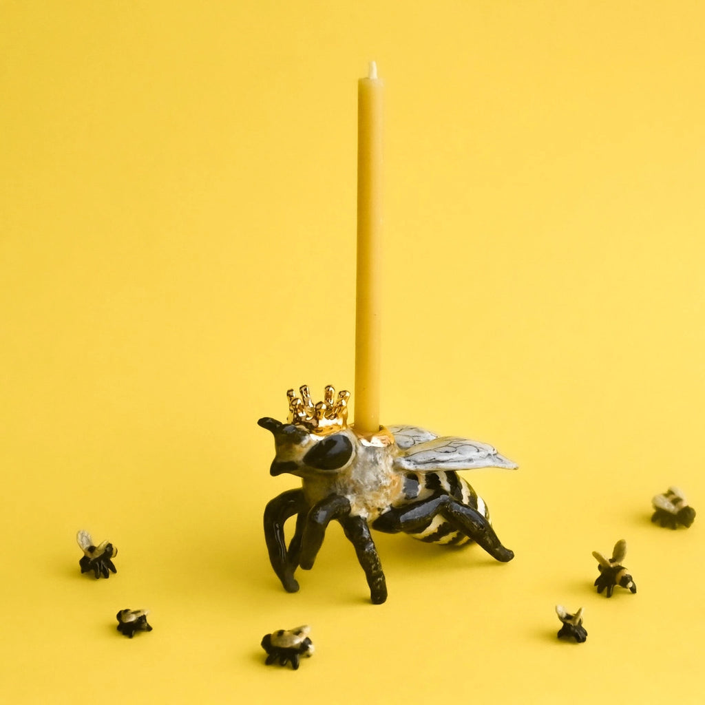 A whimsical display featuring a Queen Bee Cake Topper mounted on a fine porcelain fly sculpture adorned with a crown, surrounded by miniature flies against a bright yellow background.