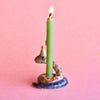 A green candle burning, inserted into a whimsical Snake Cake Topper holder shaped like a coiled snake, hand painted on fine porcelain, rests against a soft pink background.