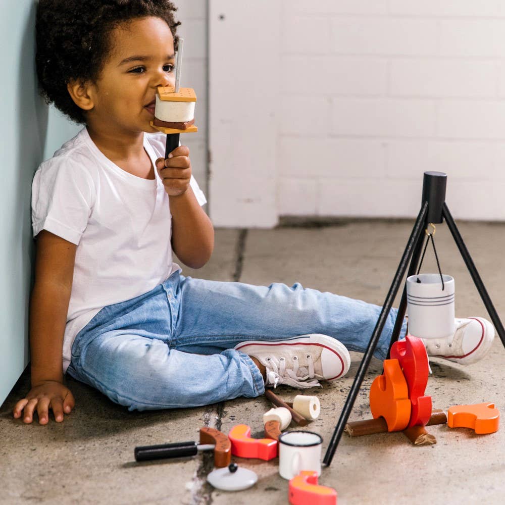 A young child enjoys a pretend s'more beside the Wooden Camp Fire Set, featuring wooden logs, marshmallows, and cups—ideal for kids' indoor camping fun. The child wears a white t-shirt, jeans, and red sneakers.