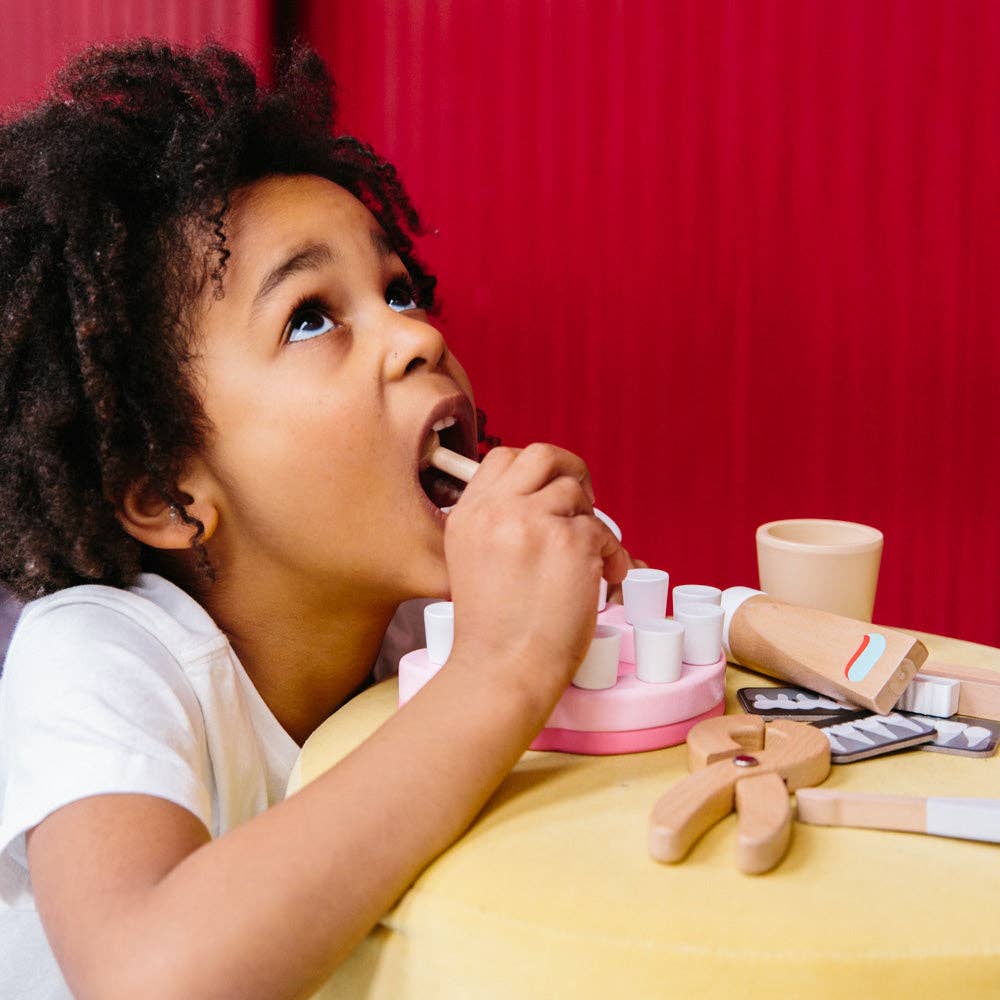 A young child plays with a Wooden Dentist Set, holding a wooden tongue depressor in their mouth at a yellow table with a red background.
