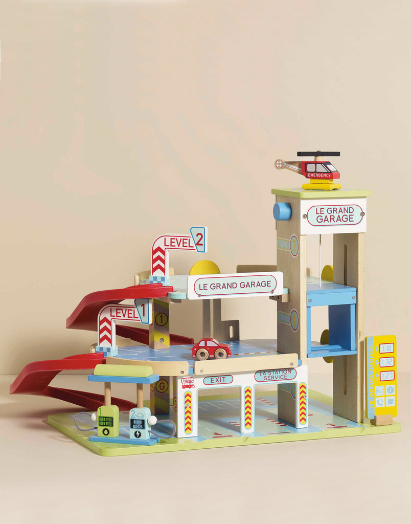 Le Grand Wooden Garage is a colorful wooden toy with three levels, ramps, an elevator, signs (“LE GRAND GARAGE,” “LEVEL 1,” “LEVEL 2”), toy cars, and a helicopter—perfect for imaginative play.