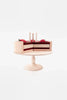 A handmade chocolate layer cake on a stand displaying a realistic model of a slice of cake, complete with decorative strawberries on top and intricate layer detailing, all crafted with non-toxic paint, set against a plain white background.