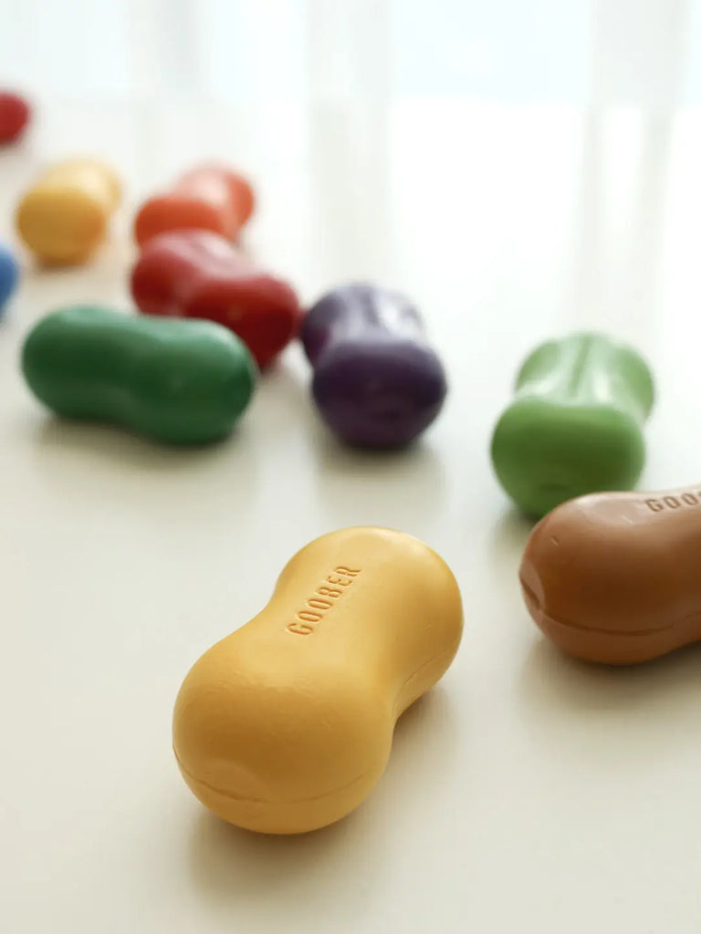 Colorful Peanut Crayons in various bright shades are scattered on a white surface, with a yellow one in sharp focus at the forefront displaying the word "goodies".