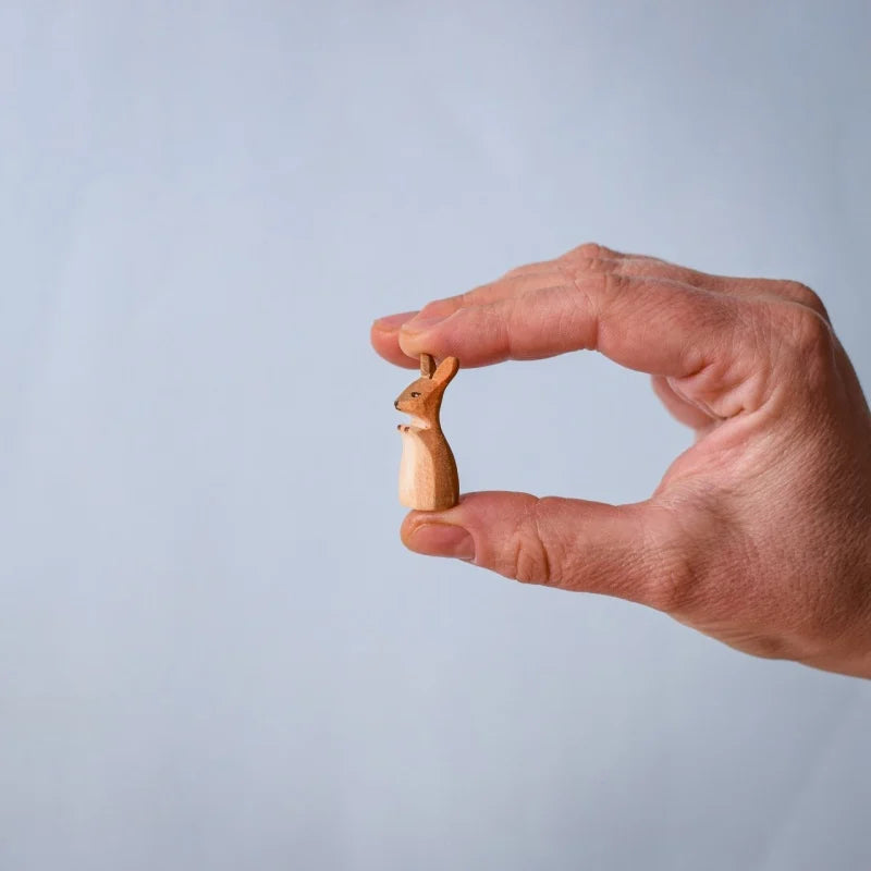 A hand holds a small kangaroo from the Bumbu Wooden Kangaroo Family Set, carved from sustainably sourced wood, upright between the thumb and index finger against a plain, light background.