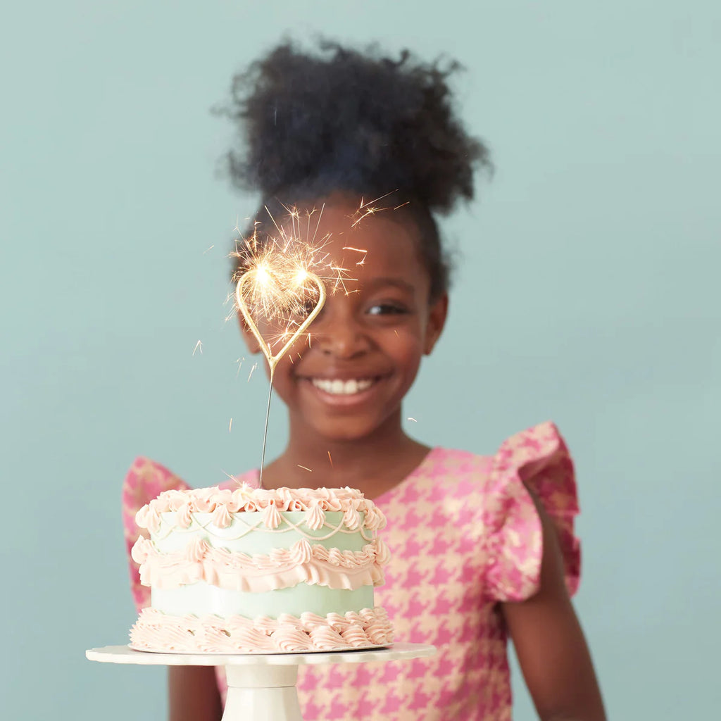 A young girl smiling beside a birthday cake with Meri Meri Gold Sparkler Heart Candles on top, against a soft blue background.
