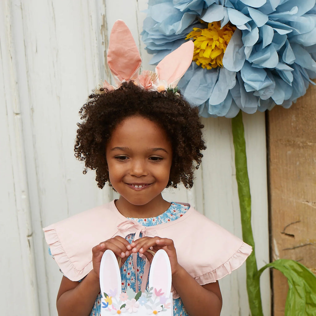 A curly-haired girl in a floral dress wears an Embellished Gingham Bunny Headband and holds a bunny-eared basket, with a large blue and yellow organza flower decorating the background.