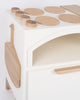 A modern white wooden Milton & Goose Play Kitchen With Pizza Oven - Made in USA partially visible in the frame, featuring a curved design with natural wood accents. The top has assorted oval and circular wood plates, and represents an interactive educational toy.