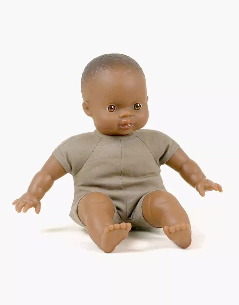 The Minikane Soft Body Doll (11") - Ondine with brown skin and short black hair is sitting upright. Dressed in a plain grey one-piece outfit, the doll has charming brown eyes. Made from phthalate-free vinyl, it makes a perfect bedtime companion. The background is plain white.