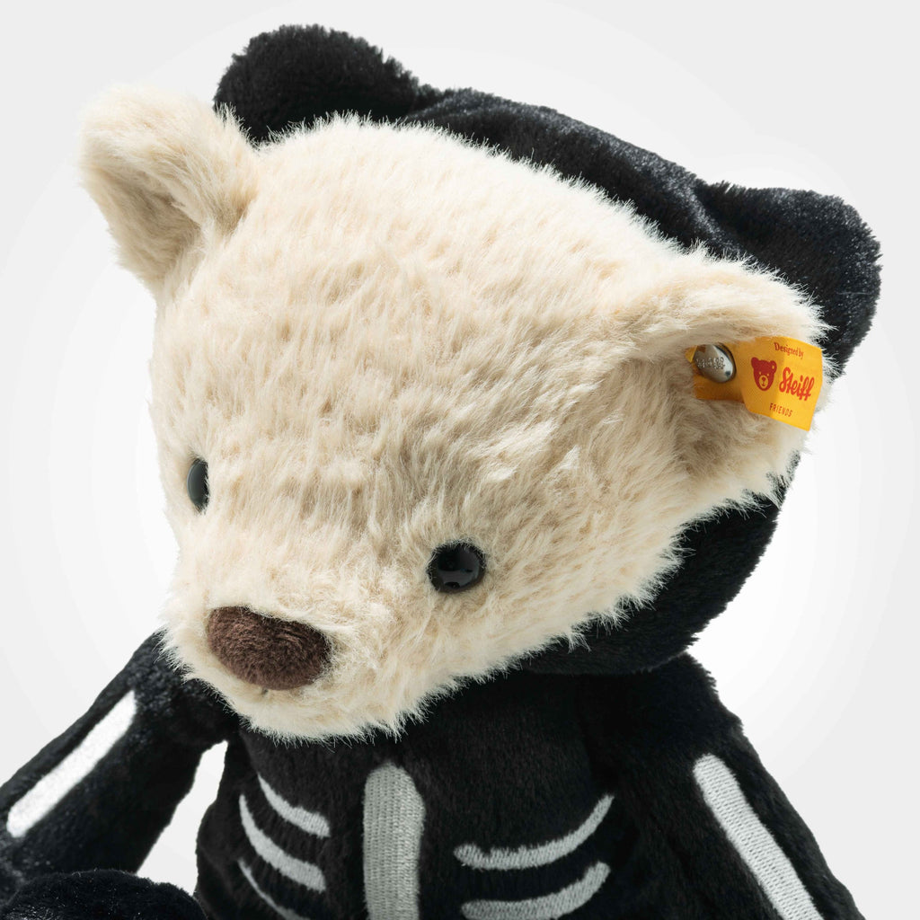 The Steiff Mr. Bones Teddy Bear features cream-colored fur, a black nose, and a skeleton costume with white details. A yellow Steiff tag in its ear makes it a standout plush for collectors, perfect for Halloween displays.