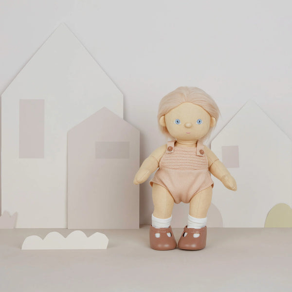 A Olli Ella | Dinkum Doll - Petal with blonde hair and blue eyes, wearing a pink outfit and brown shoes, stands in front of a simple backdrop featuring white and pink geometric shapes resembling houses.