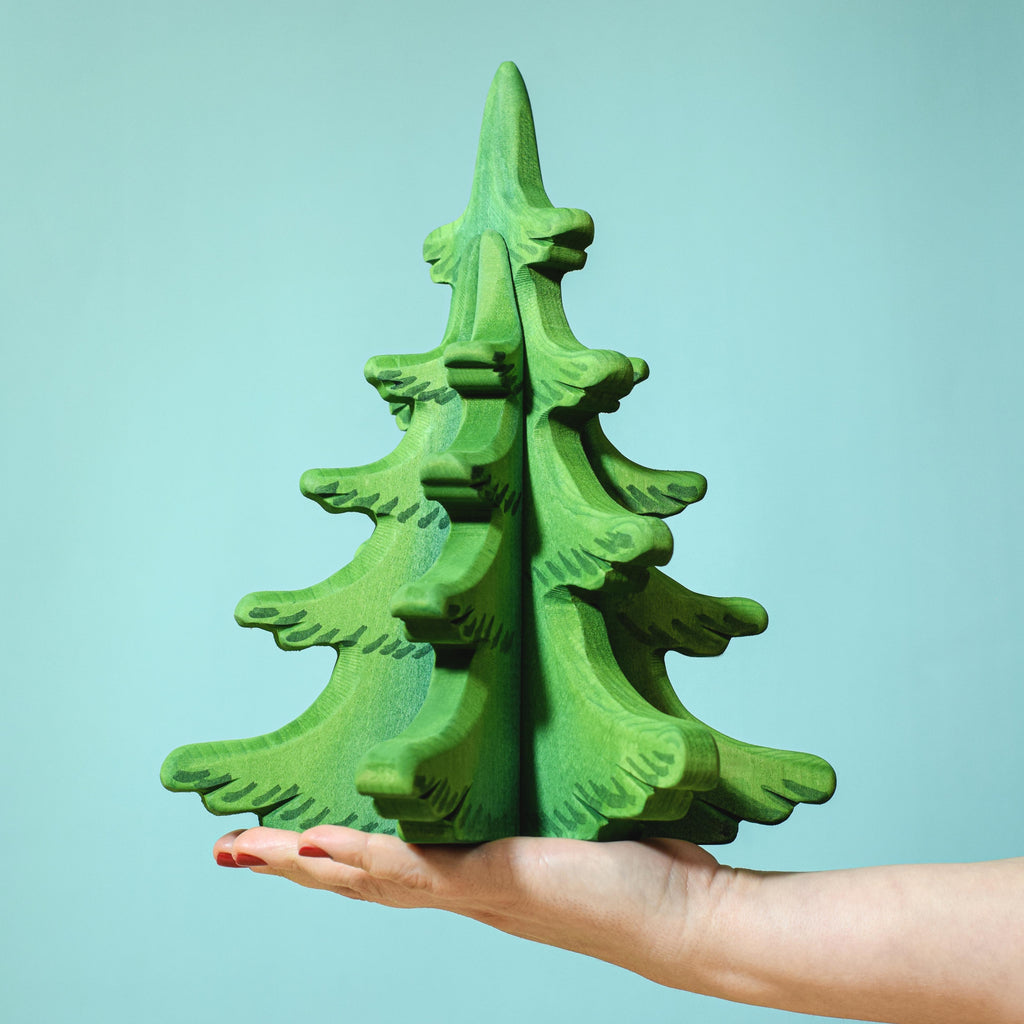 A hand with red-painted nails holding a stylized Extra Large Wooden Sugar Pine Tree decoration against a turquoise background.