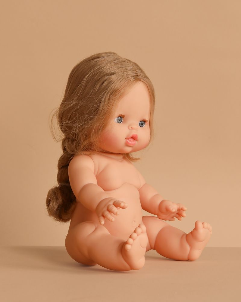 The Minikane Baby Doll (13") - Eleanor, featuring long light brown braided hair and blue eyes, sits unclothed with outstretched arms and bent legs on a beige surface against a beige background. Clothing sold separately.