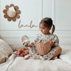 A young girl sits on a cozy bed playing with toys in a warmly decorated room, her attention focused on an Olli Ella Rattan Lily Basket Set and a plushie.