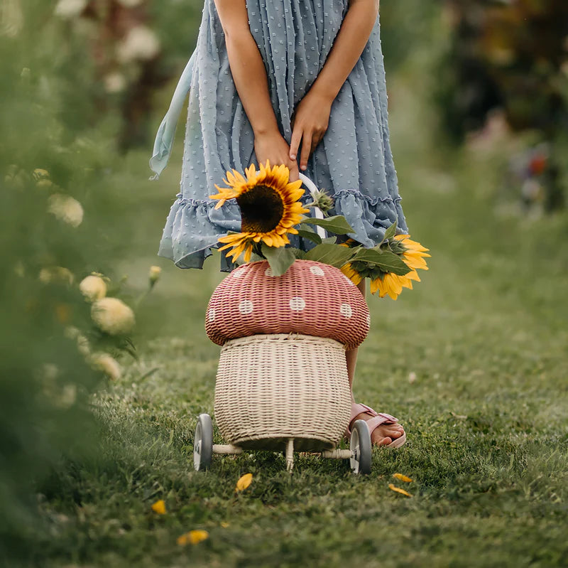A child in a blue dress stands on grass, holding sunflowers beside the Olli Ella Rattan Mushroom Luggy - Pink, a hand-woven rattan basket with a charming polka-dotted top.