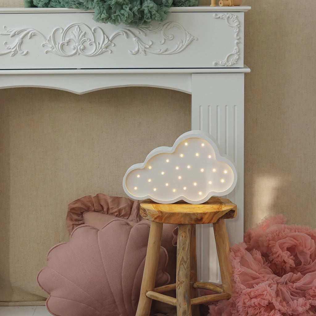 The Handmade Wooden Lamp - White Cloud, crafted from natural pinewood with small bulbs, sits on a wooden stool by a white ornate fireplace, surrounded by pink and green decorative pillows.