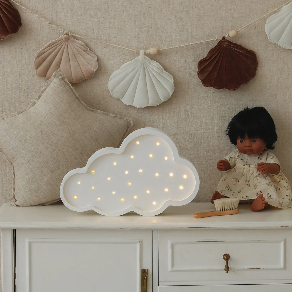 A soft toy doll in a floral dress sits on a white cabinet beside the Handmade Wooden Lamp - White Cloud, with a star-shaped pillow and a fabric seashell garland hanging on the wall.