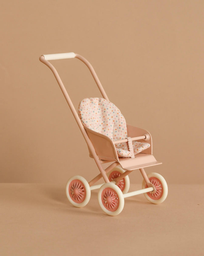 The Maileg Stroller, Micro - Powder is a small, pastel pink stroller with white wheels and a polka-dot fabric seat, shown against a beige background.
