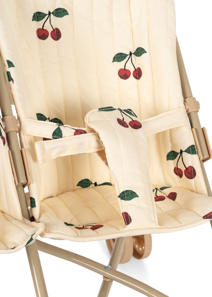 A close-up of the Konges Sloejd Twin Doll Stroller - Cherry shows its padded cream-colored seats with red cherry and green leaf patterns, safety straps, and a light beige metal frame.