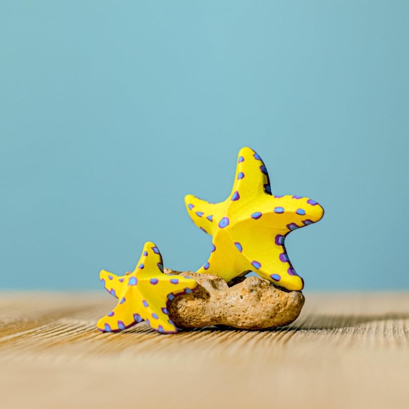 The Bumbu Handmade Wooden Yellow Starfish Set features two yellow wooden starfish with purple spots on a light brown rock, set against a wooden surface and blue background—an eco-friendly Montessori educational toy.