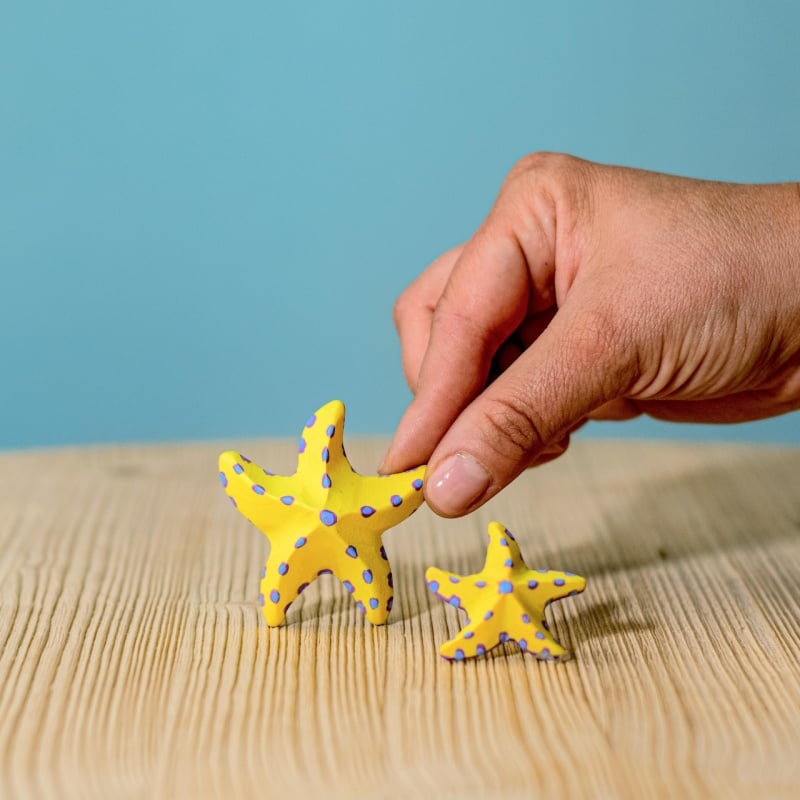 A hand holds a small starfish from the Bumbu Handmade Wooden Yellow Starfish Set, featuring yellow wood with blue spots beside a matching larger piece on a light wooden surface and blue background—ideal for Montessori learning.