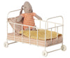 A Maileg Micro Cot bed with wheels, covered with a pink floral blanket and a yellow knit blanket, with a stuffed rabbit toy sitting inside.