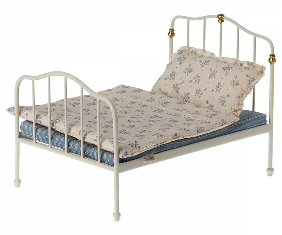 A Maileg Bed, Parent Mouse - Off White with a white frame featuring delicate, curved designs and small decorative knobs at the headboard and footboard. Dressed in Maileg printed fabrics, the bed boasts a blue-striped mattress, a floral comforter, and matching pillows adorned with light blue flowers—perfect as a bed for bigger mice.