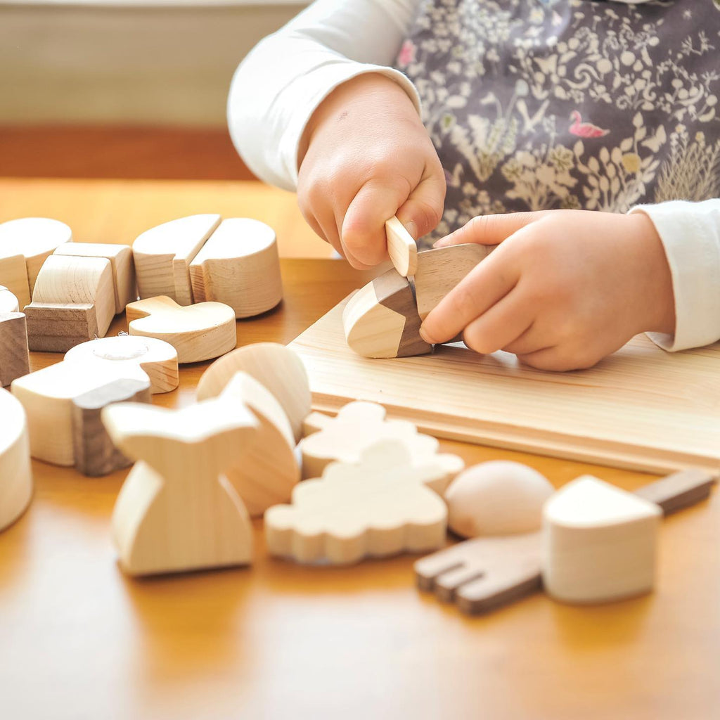 A child's hands playing with various wooden shapes including animals, geometric blocks, and Velcro Wooden Vegetables on a wooden table, focusing on fitting pieces together.