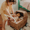 A young child in a yellow outfit lovingly adjusts a doll in an Olli Ella Rattan Doll Stroller, standing on a cozy rug in a warmly lit bedroom.