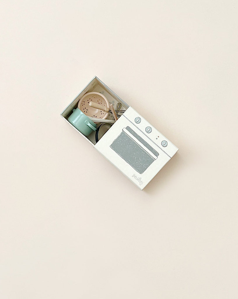 A minimalist image of a Maileg Cooking Set in a playful stove box against a pale pink background, including a stove, a sink, and kitchen utensils.