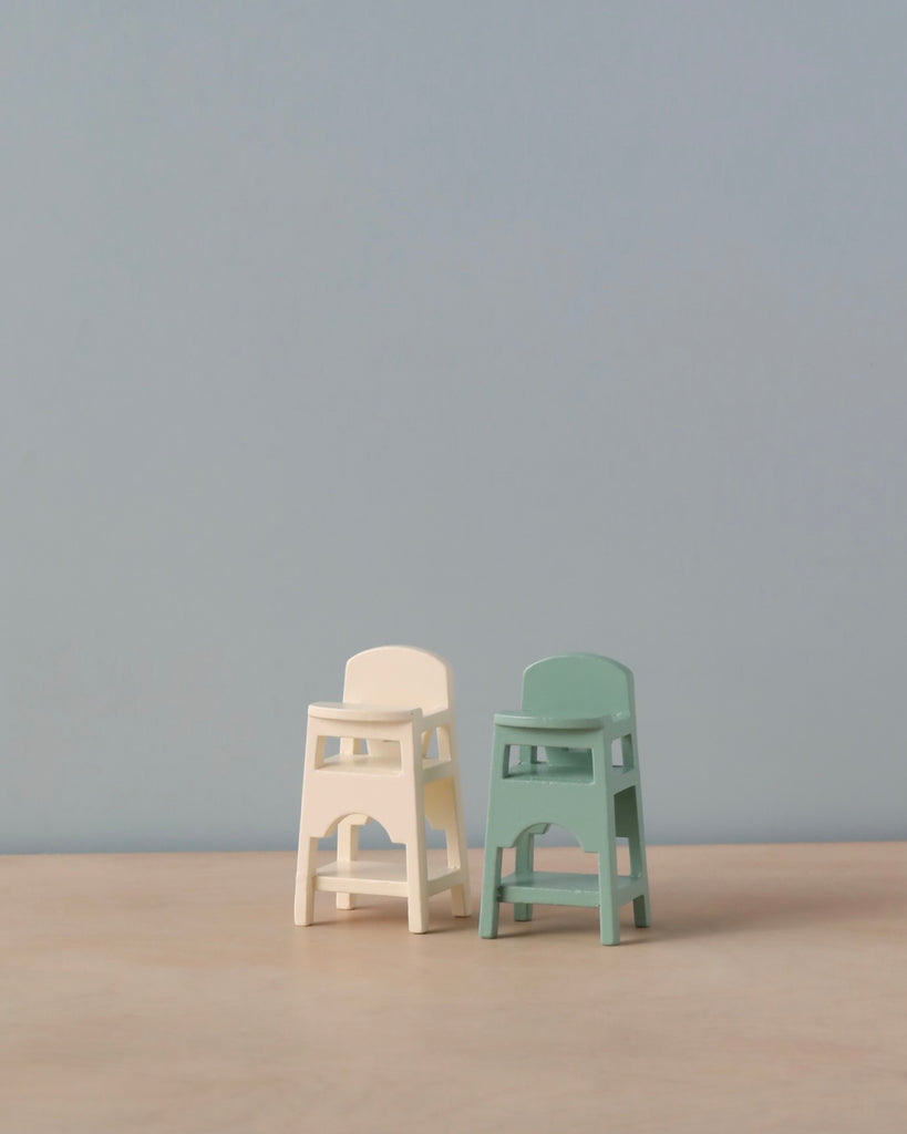 Two small Maileg Mouse Size High Chairs, one white and one green, positioned on a wooden surface against a light blue background, suggesting simplicity and minimalism.