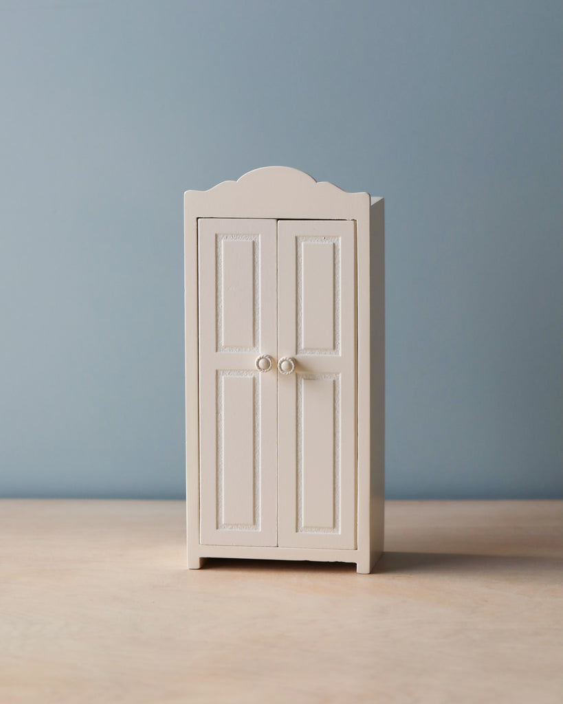 A Maileg Small Closet with double doors and round knobs stands on a light wooden surface against a plain, blue-gray background. Made of FSC wood, the closet boasts a simple and elegant design with decorative molding along the top edge and door panels. Inside, golden hangers gleam invitingly.