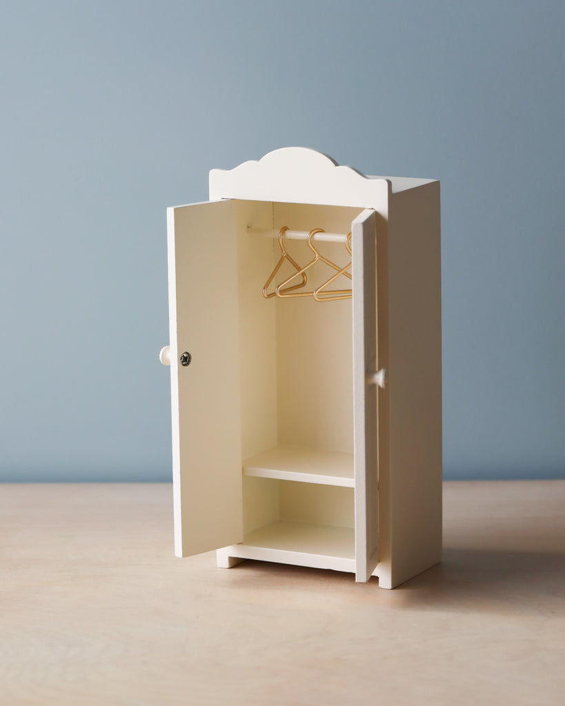 A Maileg Small Closet made of FSC wood with one door partially open, revealing two golden hangers inside. The closet stands on a light wood surface with a light blue background.
