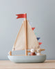 A Wooden Sail Boat with beige fabric sails on a blue-gray base, decorated with red and multicolored flags. Two cartoonish, animal-like figures are on the boat against a plain light background