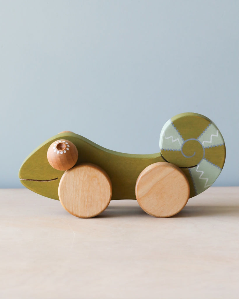 Wooden green chameleon with light blue stripes on tail, natural color wheels and eyes. Beige counter and light blue background.