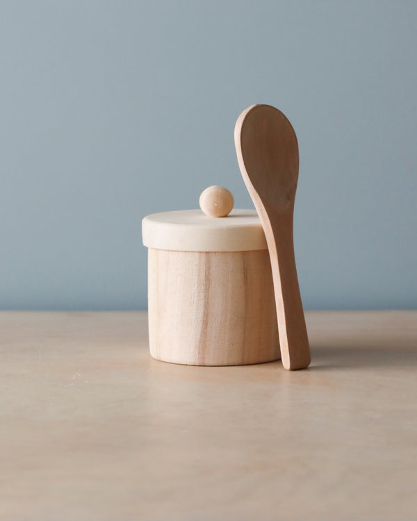 A simple Wooden Doll Feeding Set with a round lid and a small spherical handle, paired with a wooden spoon, set against a pale blue background.