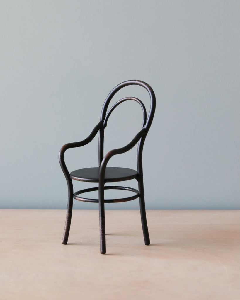 A minimalistic photo of a single Maileg | Chair with Armrest, depicted with a round back, placed centrally on a wooden floor against a light grey background.