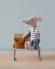 A Maileg Big Brother With Backpack toy in a striped shirt and blue jeans sits on a small wooden bench, holding a tiny brown school backpack, against a soft blue background.