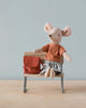 A small Maileg Big Sister With Backpack - Old Rose wearing an orange striped shirt and blue plaid shorts sits on a miniature wooden bench. Next to the mouse is a tiny red handbag. The background is light blue, and the bench rests on a smooth light wood surface, evoking charm reminiscent of kindergarten toys.