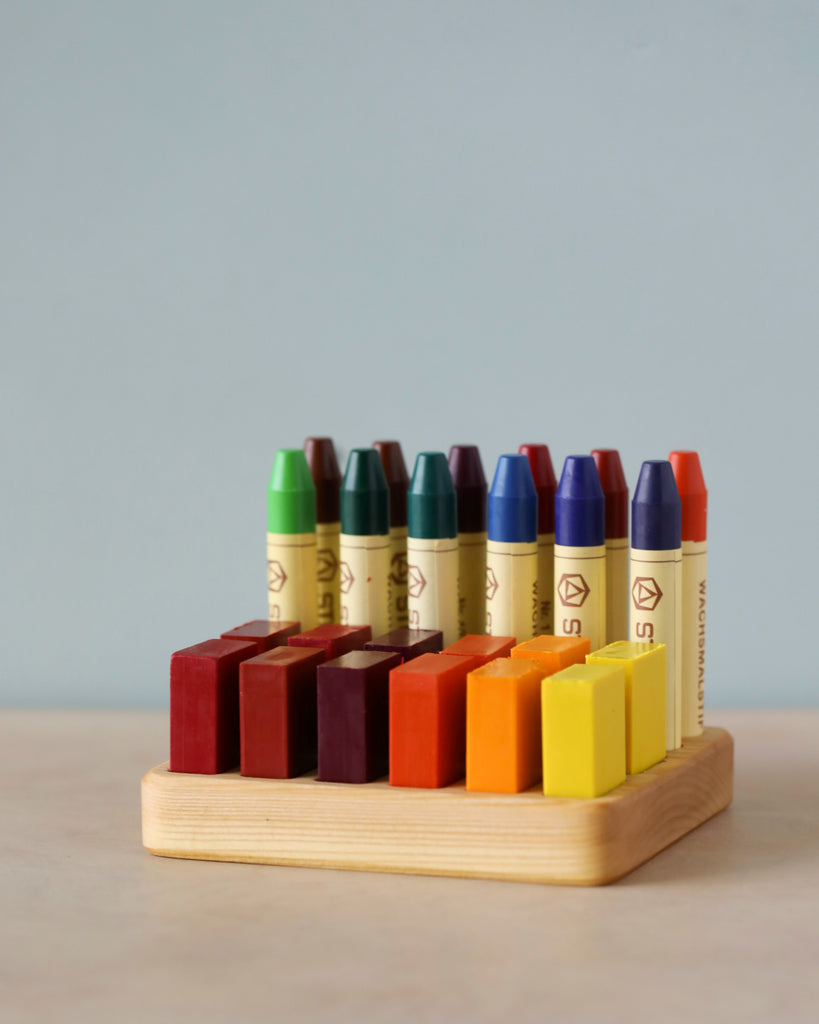 A row of colorful Crayon Tray For Stockmar -12 x 12 Slots in a Linden wood holder against a light blue background. The crayons are neatly arranged in a gradient, transitioning from green to orange.