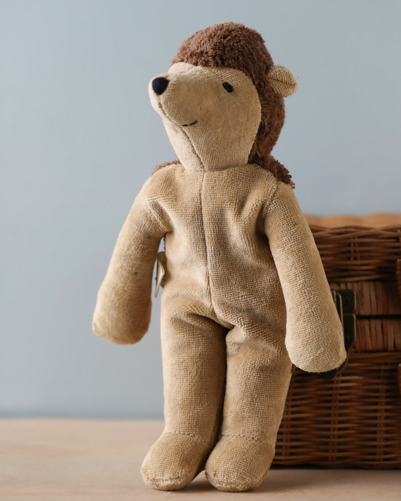 A Senger Naturwelt stuffed animal hedgehog wearing a zip-up onesie made from organically grown cotton with a hood shaped like a monkey's head, sitting against a wicker basket on a plain background.