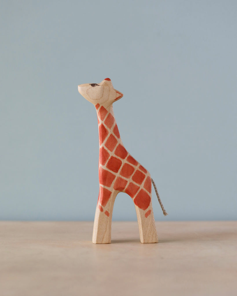 A Handmade Holzwald Giraffe - Small figurine with painted red patches and a visible grain texture, standing against a light blue background. This high-quality toy adds educational value and aesthetic appeal to any collection.