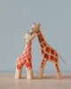 Two Handmade Holzwald giraffe figurines, one large and one small, touching noses affectionately on a soft blue background. The giraffes, sustainable toys made from wood, are patterned with red and
