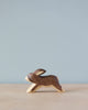 A small wooden Handmade Holzwald Running Rabbit figurine on a plain surface with a soft blue background. The rabbit is stylized and simplistic, with visible wood grain.