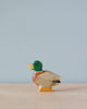 A small, Handmade Holzwald Male Duck figurine with a green head and natural wood body stands against a soft blue background.