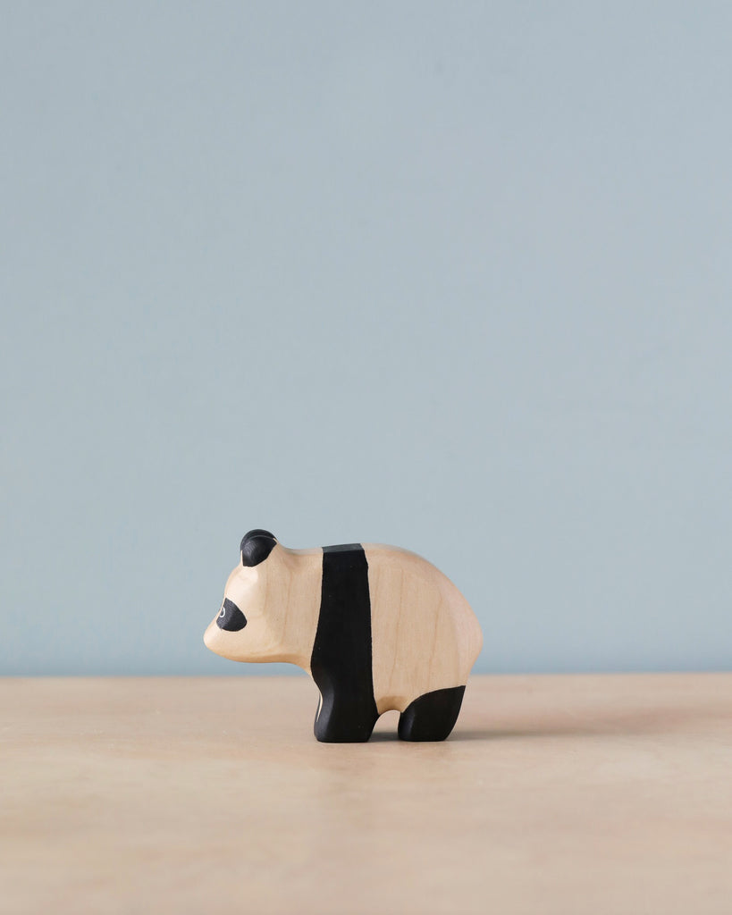 A handmade Holzwald Baby Panda, painted in natural wood and black colors, standing against a pale blue background.