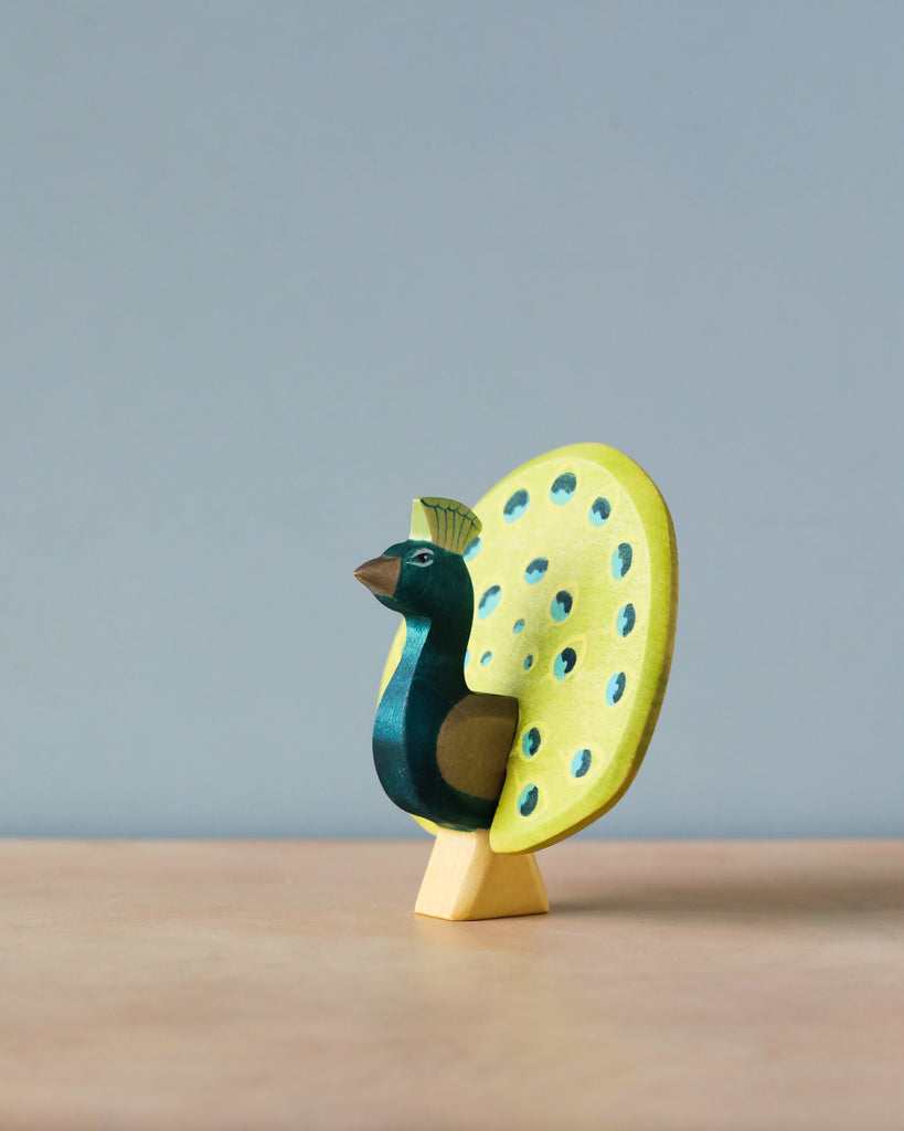 A colorful Handmade Holzwald peacock figurine with a dark blue body and light blue speckled tail on a sustainable wooden stand, set against a neutral background.