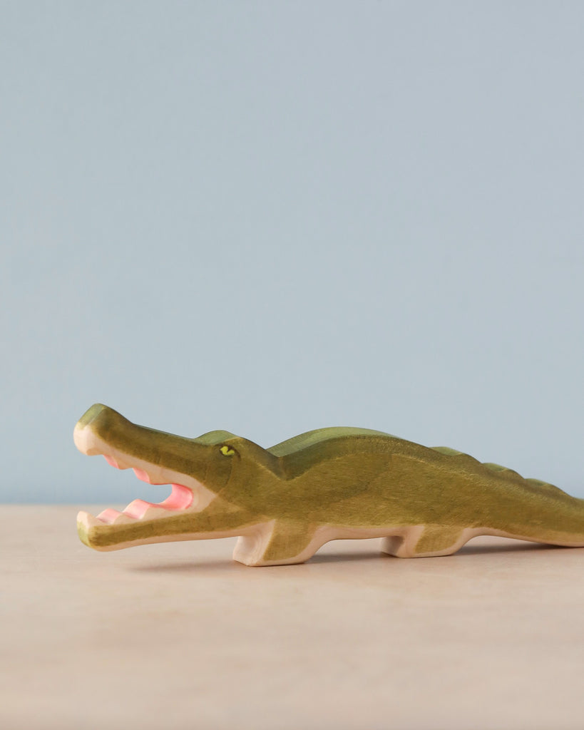 A Handmade Holzwald Crocodile with an open mouth, painted green and cream, positioned on a plain surface against a pale blue background.