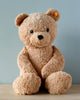 A Steiff XL Jimmy Teddy Bear with light brown fur is sitting against a pale blue background. The bear has dark, shiny eyes, a small nose, and a faint smile. A yellow tag is visible on the bear.